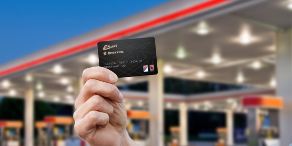 A hand holding up a black Flyers and Quick Fuel fleet card in front of a fueling station.